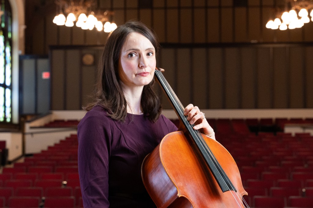 A cellist sits in a concert hall holding a cello, wearing a purple dress, with empty red seats in the background.